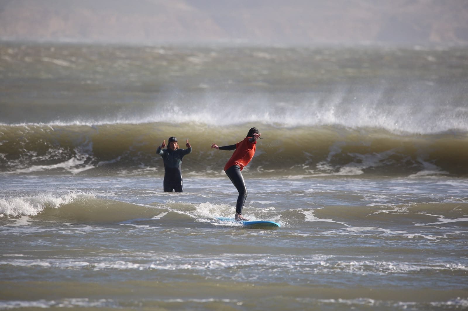 A surfer riding a small wave while the coach stands in the water giving two thumbs up