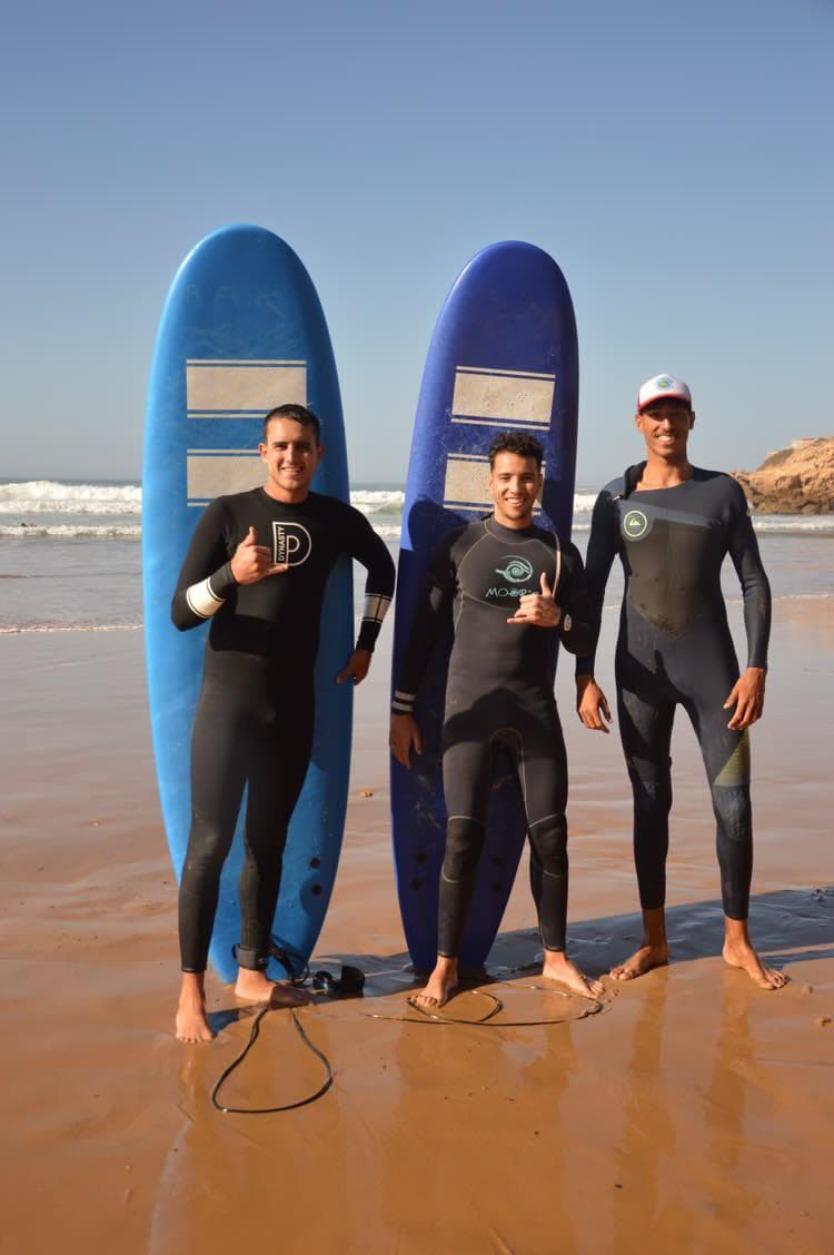Three surfers in wetsuits standing with their boards on the wet sand, making shaka signs