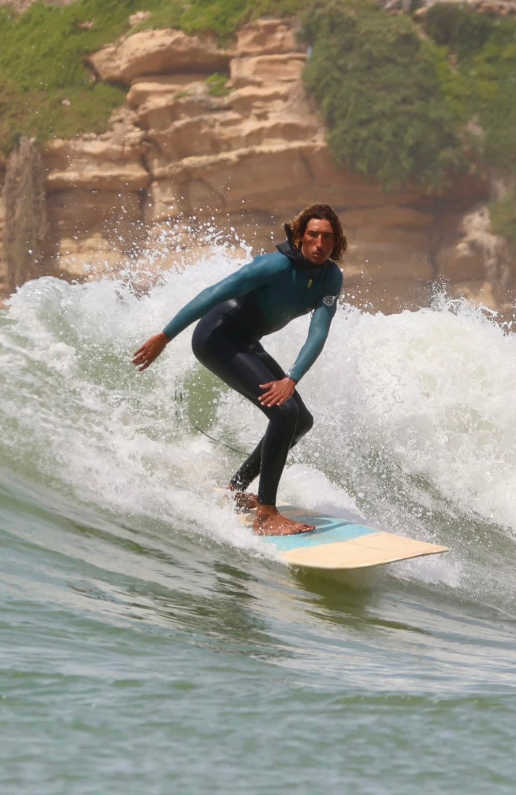 A surfer in a teal wetsuit riding along the face of a wave, crouched with one hand trailing