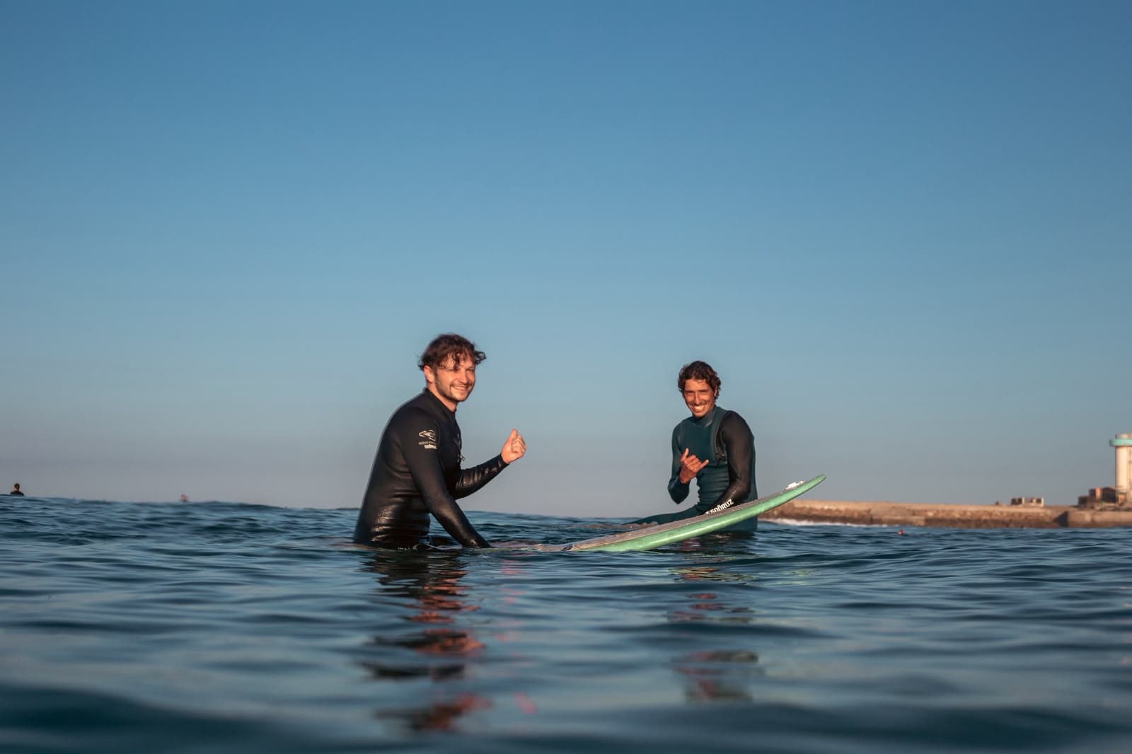 Two surfers in wetsuits sitting on their boards in calm water, smiling, with the harbour wall behind them