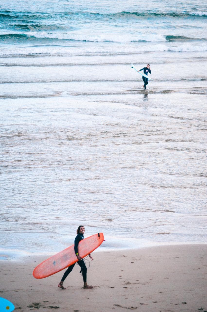 A surfer carrying an orange longboard along the shoreline as another walks out into the water